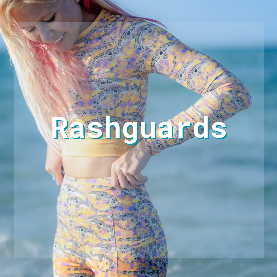 Woman standing in front of a body of water wearing a cropped long sleeve rashguard and leggings. Text over the image that says “rashguards” to indicate the items of this collection.