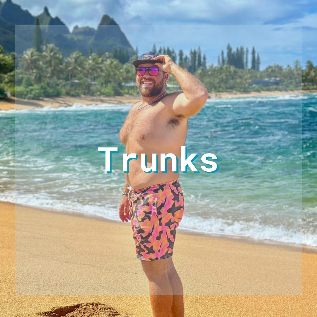 A man standing on the beach smiling wearing shark swim trunks. Text over the image says “trunks” to indicate the items in this collection. 