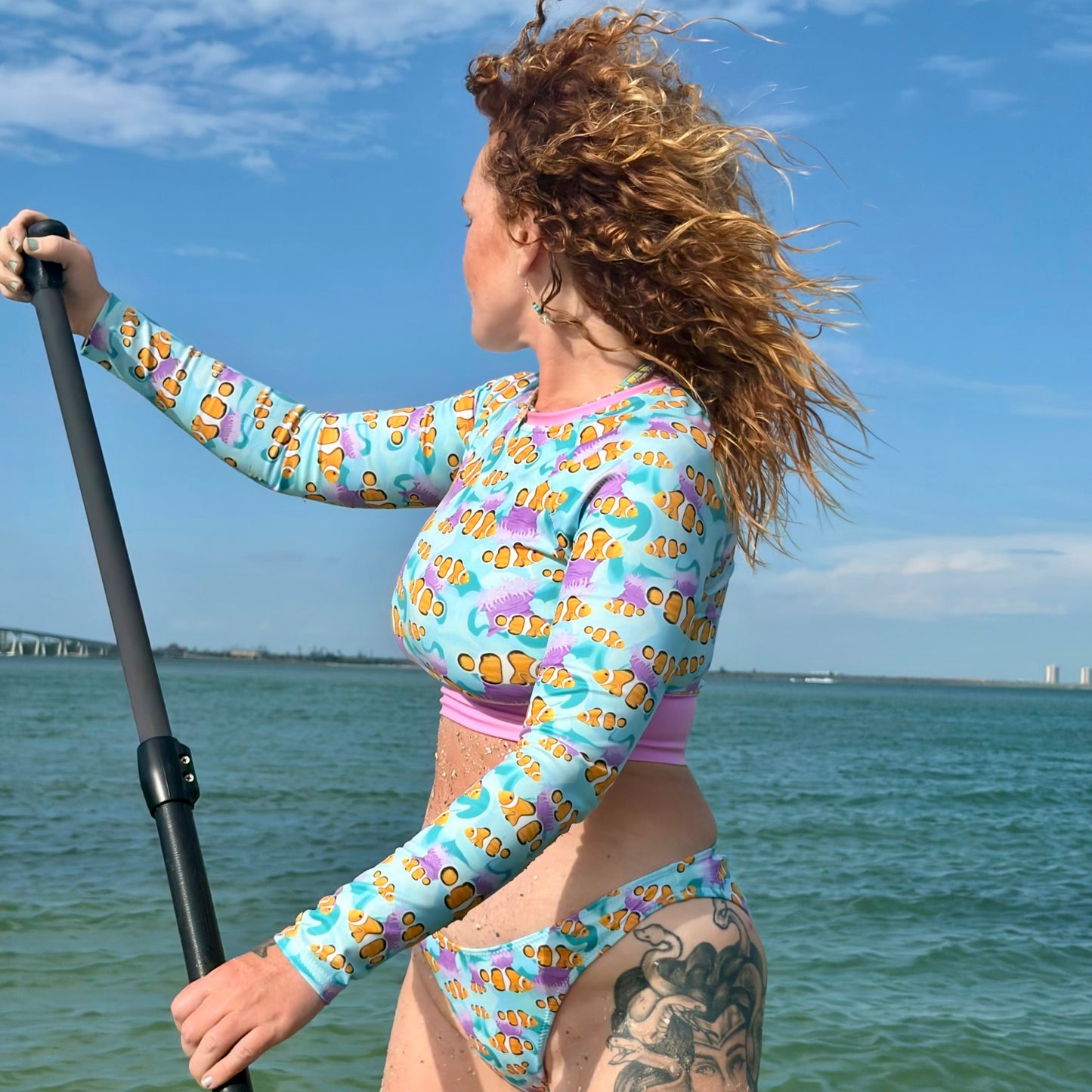 Woman paddlebording wearing a blue, pink and orange clownfish cropped rashguard and bikini bottoms 