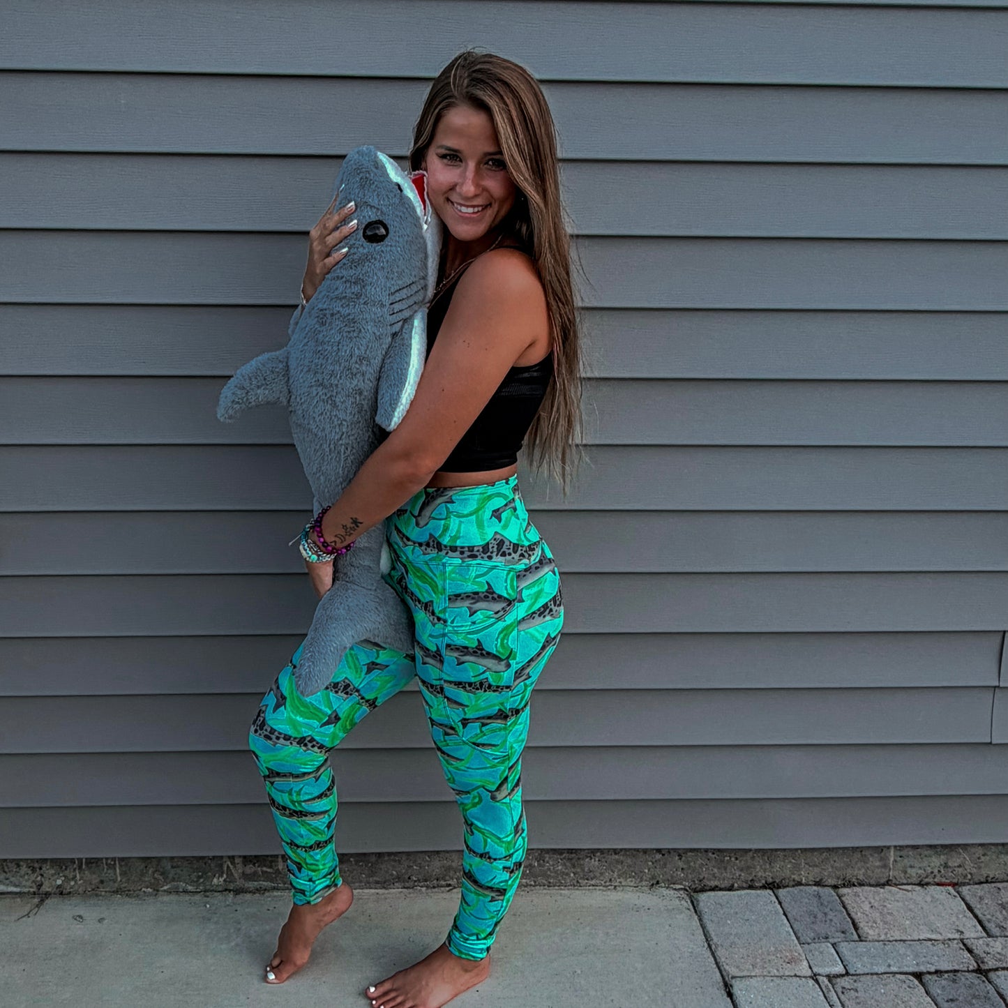 woman standing with a shark stuffed animals wearing a long sleeve green/teal rashguard and leggings with leopard sharks on it in front a grey house 