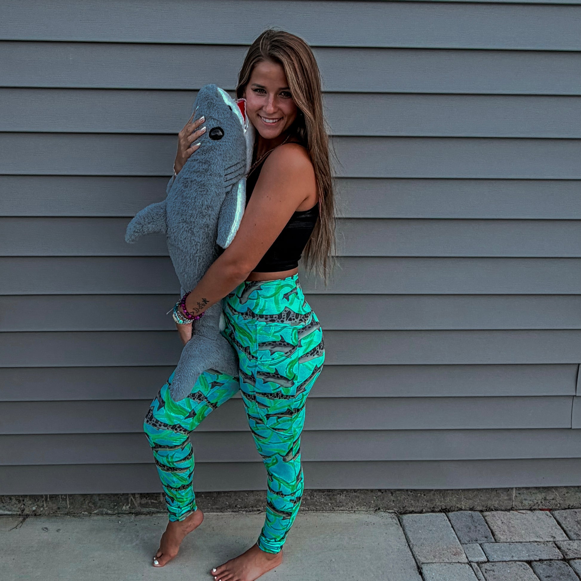 woman standing with a shark stuffed animals wearing a long sleeve green/teal rashguard and leggings with leopard sharks on it in front a grey house 