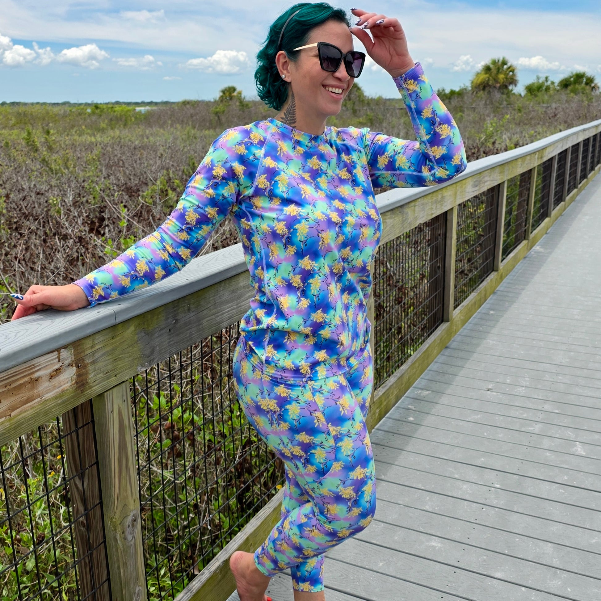 woman on a boardwalk wearing a long sleeved rashguard that is purple, green, blue, yellow with sea dragons on it 