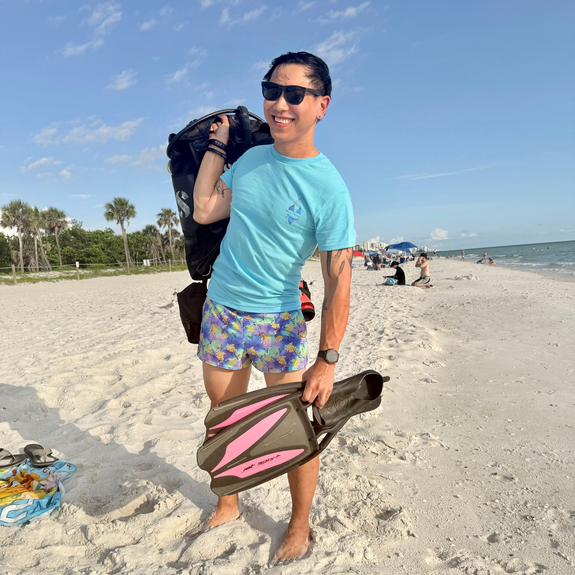 man kneeling on the beach with scuba gear wearing a blue t shirt with a logo on the front chest 