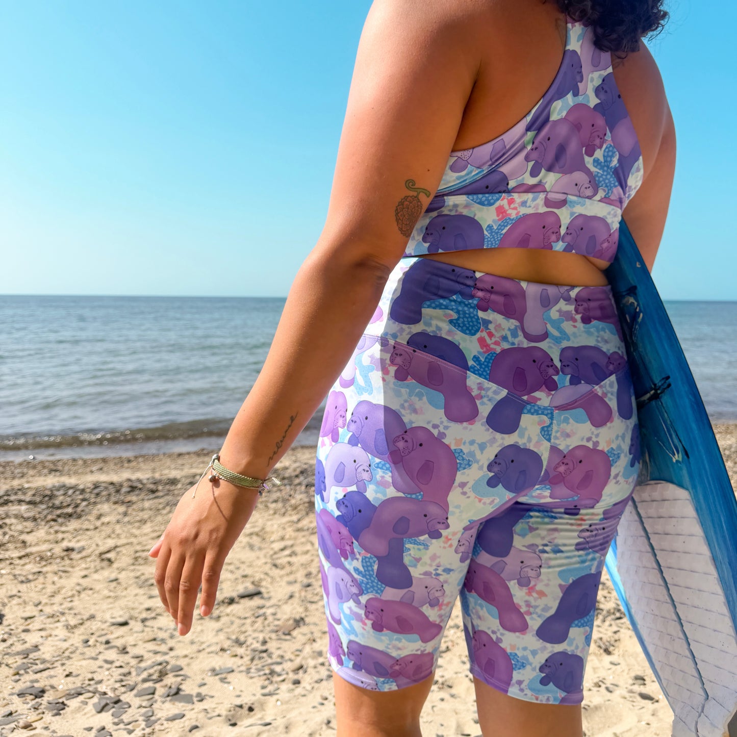 woman standing on a beach with a surfboard wearing a purple and blue manatee activewear set