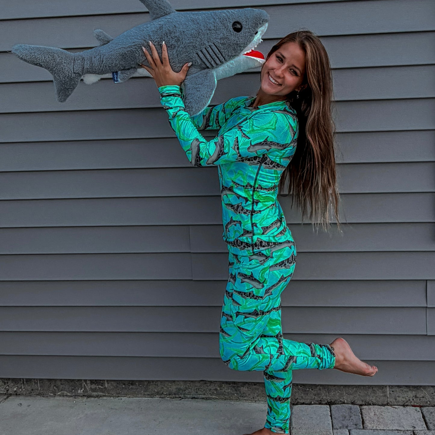 woman standing with a shark stuffed animals wearing a long sleeve green/teal rashguard and leggings with leopard sharks on it in front a grey house 