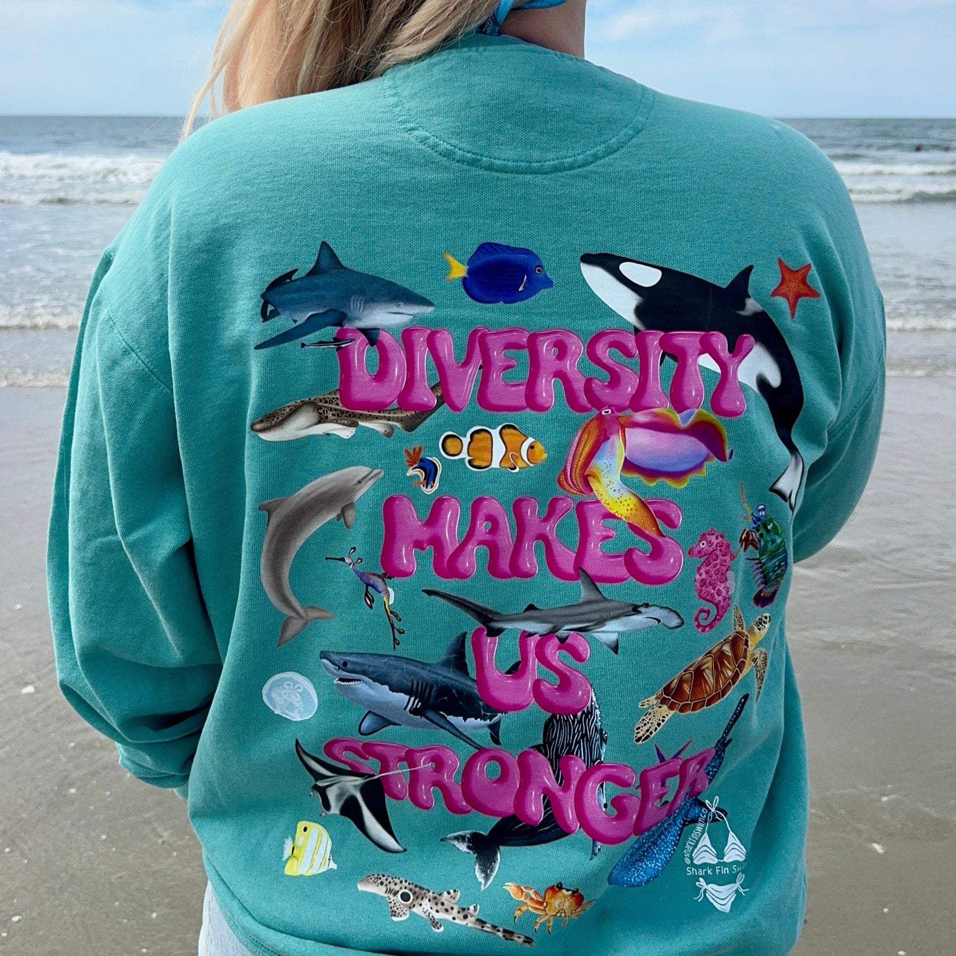 Woman on the beach wearing Green Crewneck sweatshirt with colorful ocean animal design on the back that says “diversity makes us stronger”