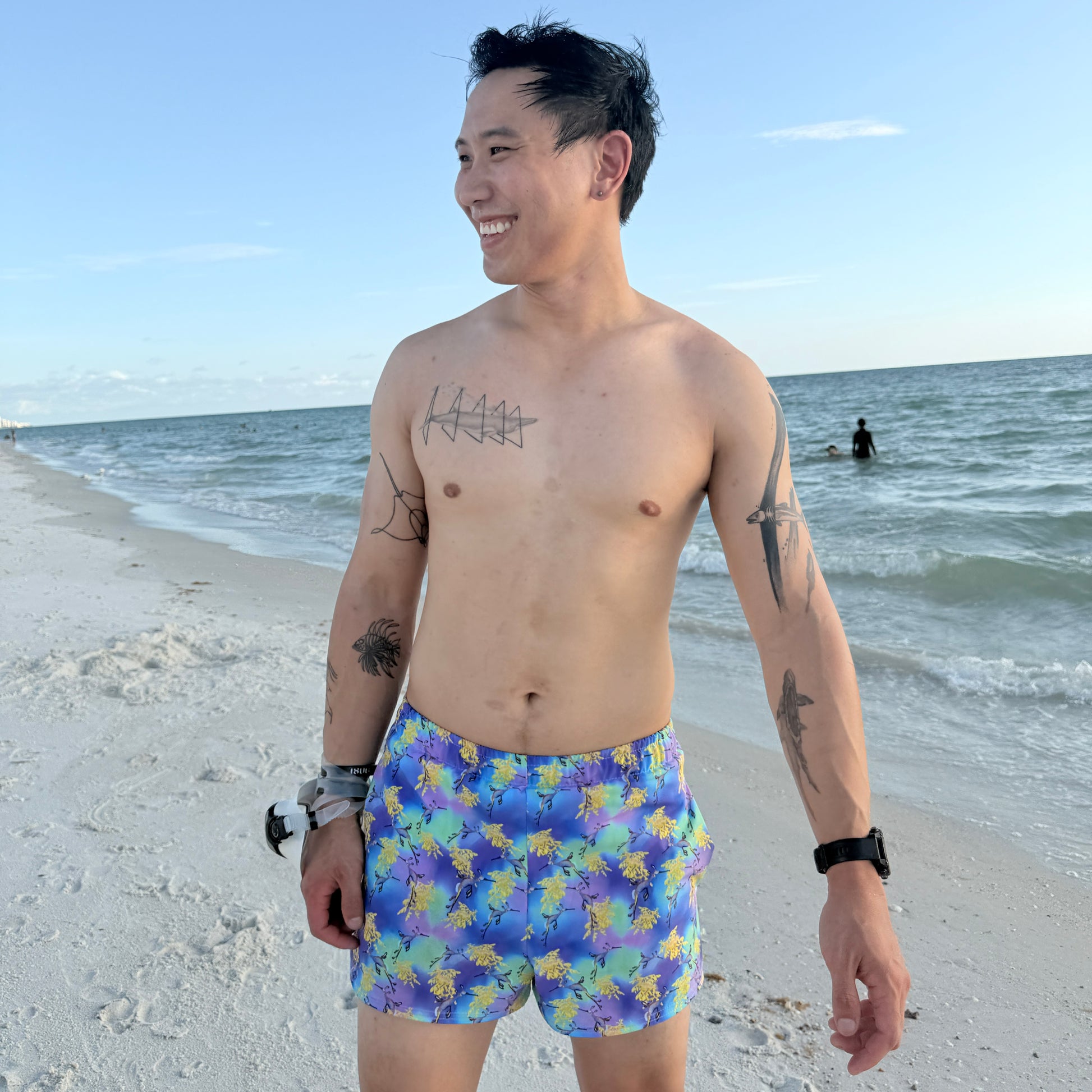 man standing on a beach by water wearing green, purple, yellow and blue swim shorts with sea dragons on them 