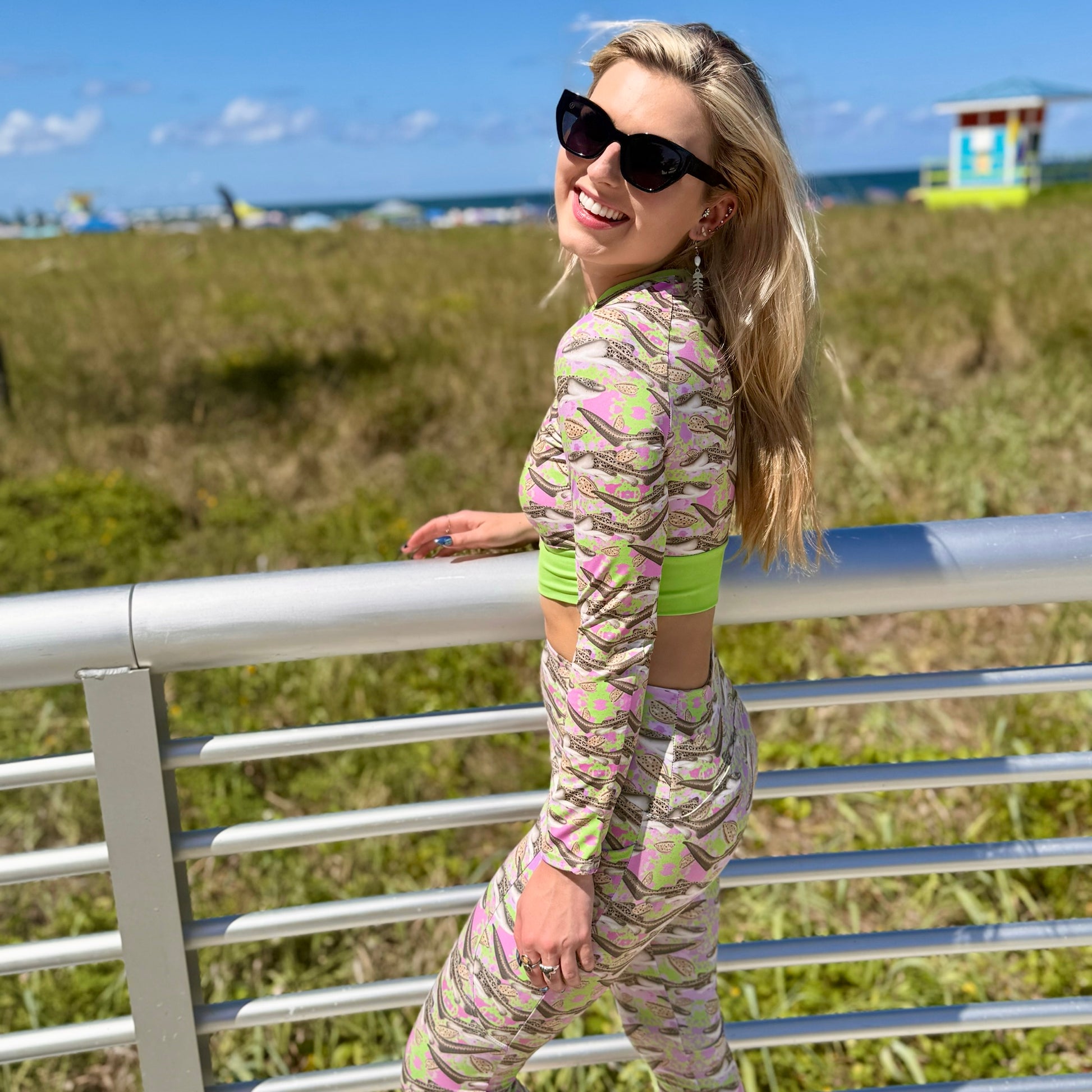 woman standing on a boardwalk by the ocean wearing a green and pink cropped rashguard and leggings with zebra sharks on it. 