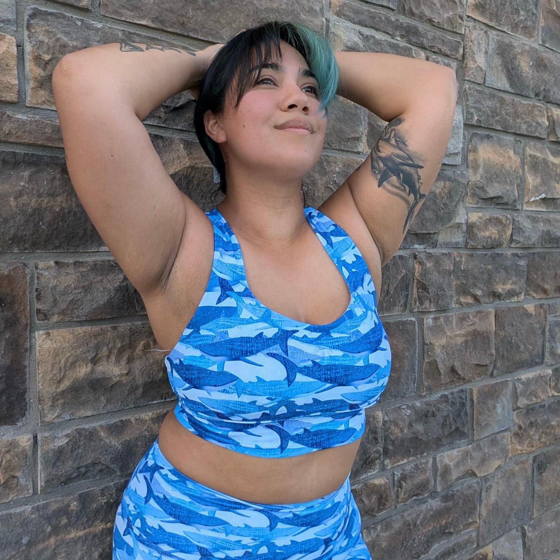 woman with black and blue hair wearing blue whale shark sports bra and active shorts against a stone wall 