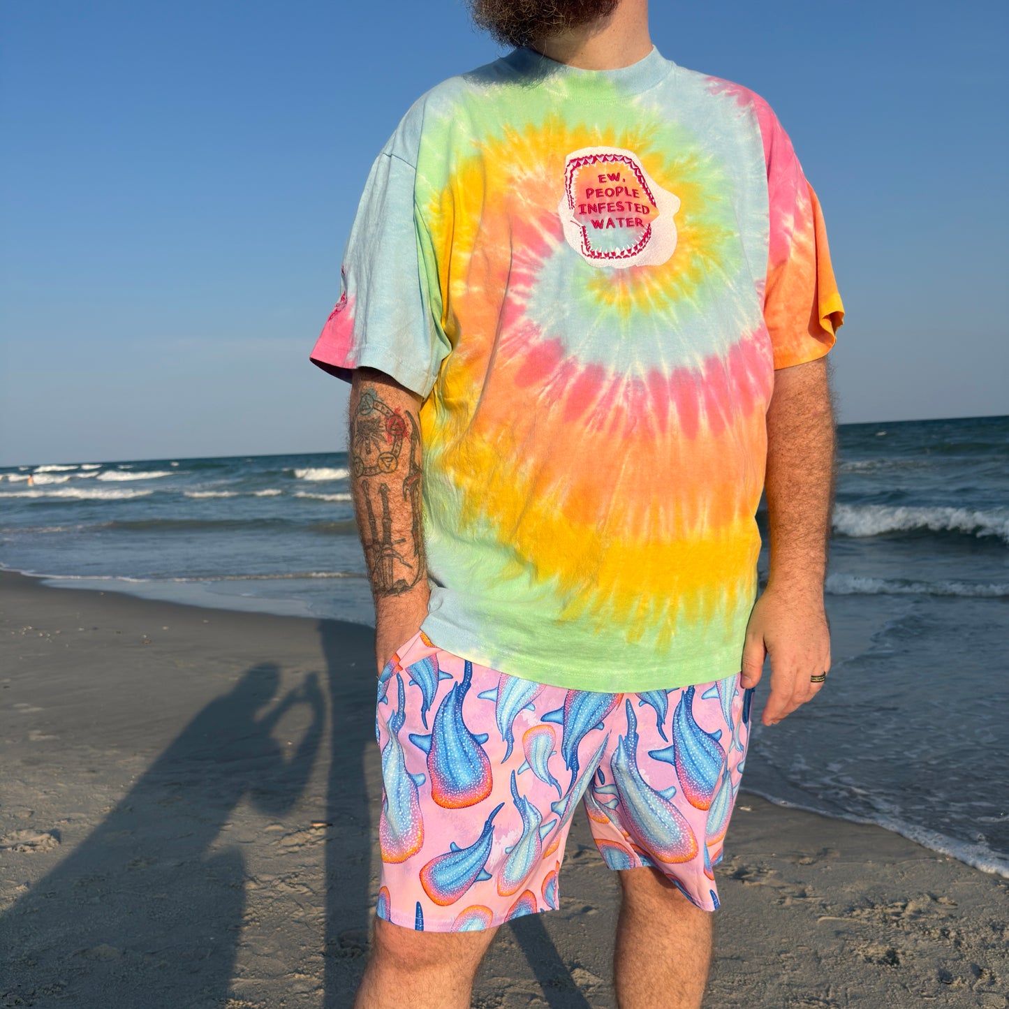 person standing on a beach by water wearing a rainbow tie dye shirt with a shark jaw and pink colorful whale shark trunks 