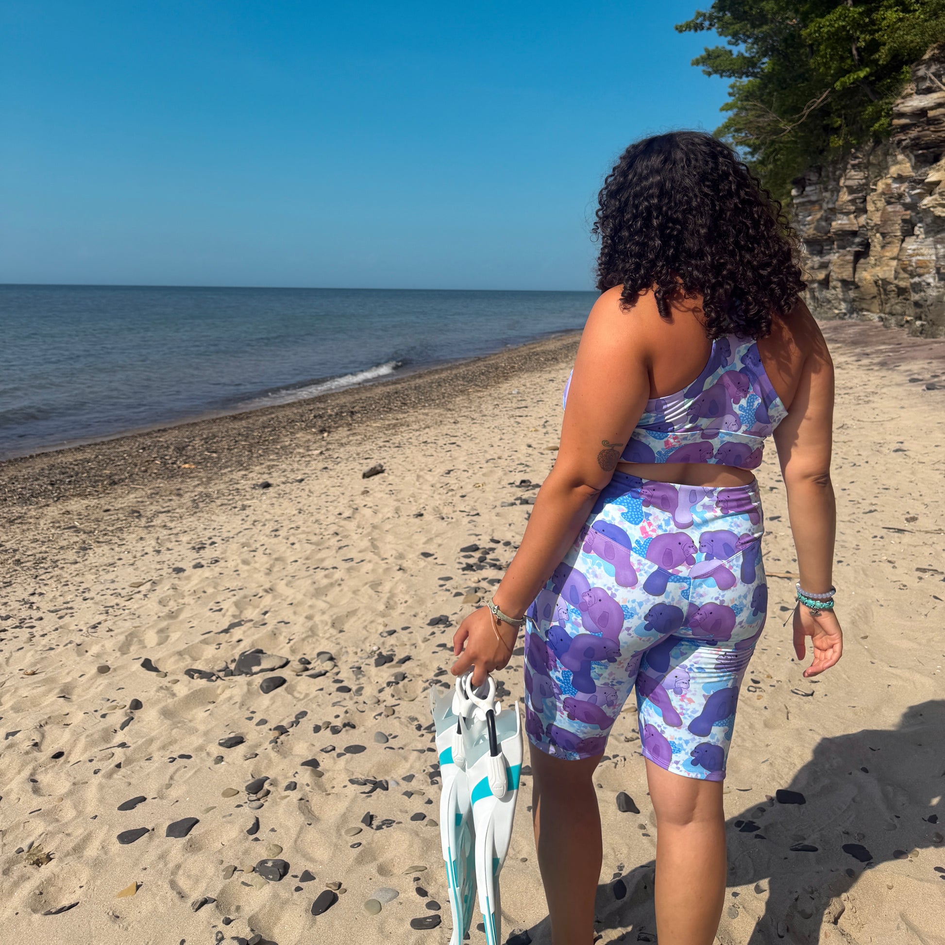 woman walking on a beach holding dive find wearing a purple and blue manatee activewear set facing away from the camera 