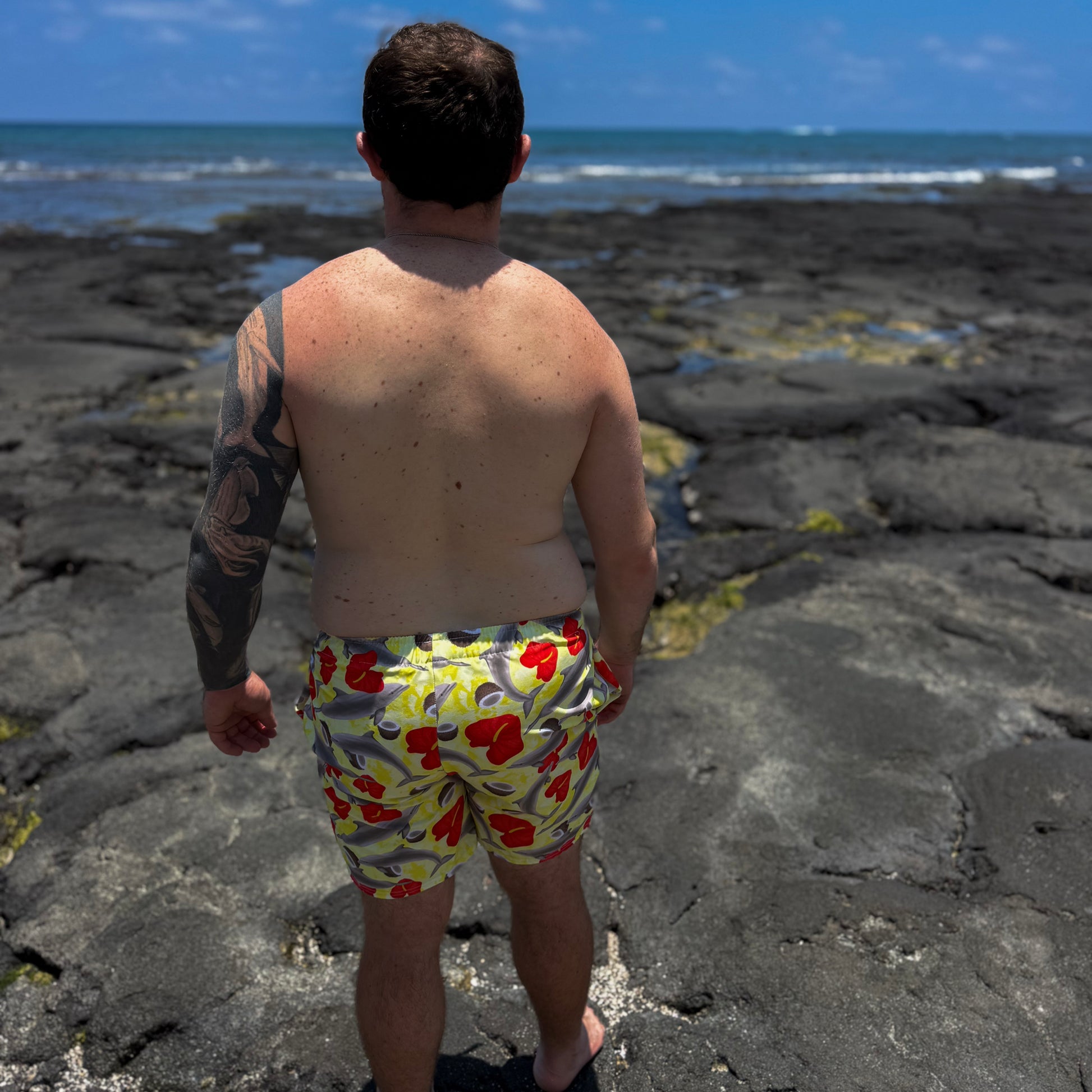 Man wearing colorful yellow and red dolphin swim trunks standing on a rocky beach with ocean in the background