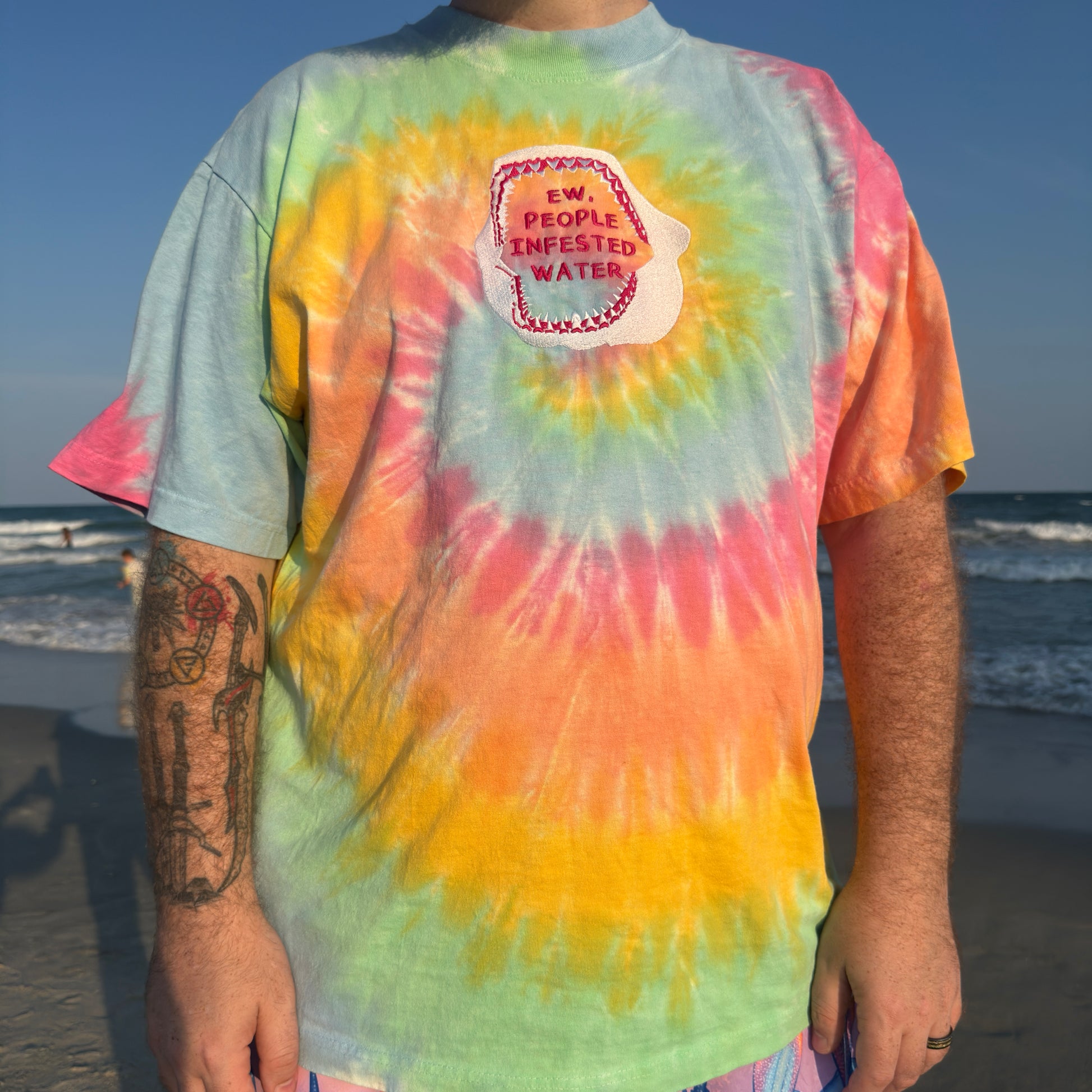 person standing on a sandy beach wearing a rainbow tie dye shirt with a shark jaw embroidered on it with text inside 