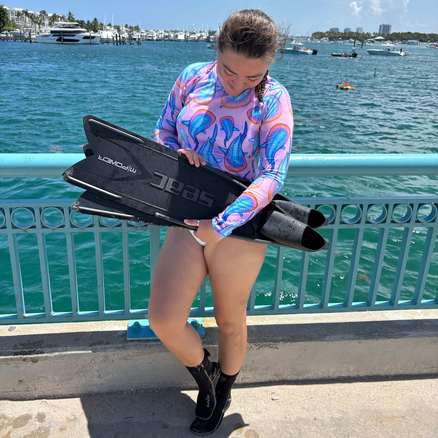 woman standing in front of a metal rail by water wearing a long sleeve pink and colorful whale shark rashguard holding dive fins 