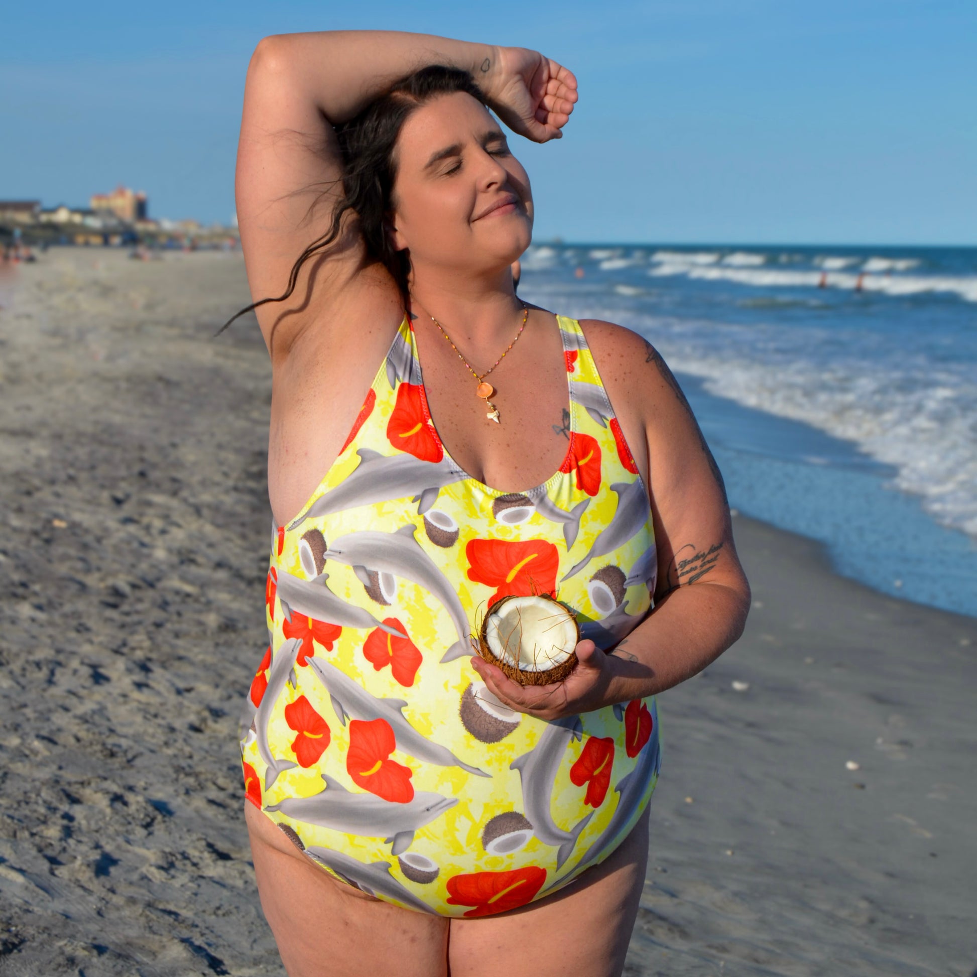 Woman on the beach wearing yellow and red dolphin one piece swimsuit holding a coconut 