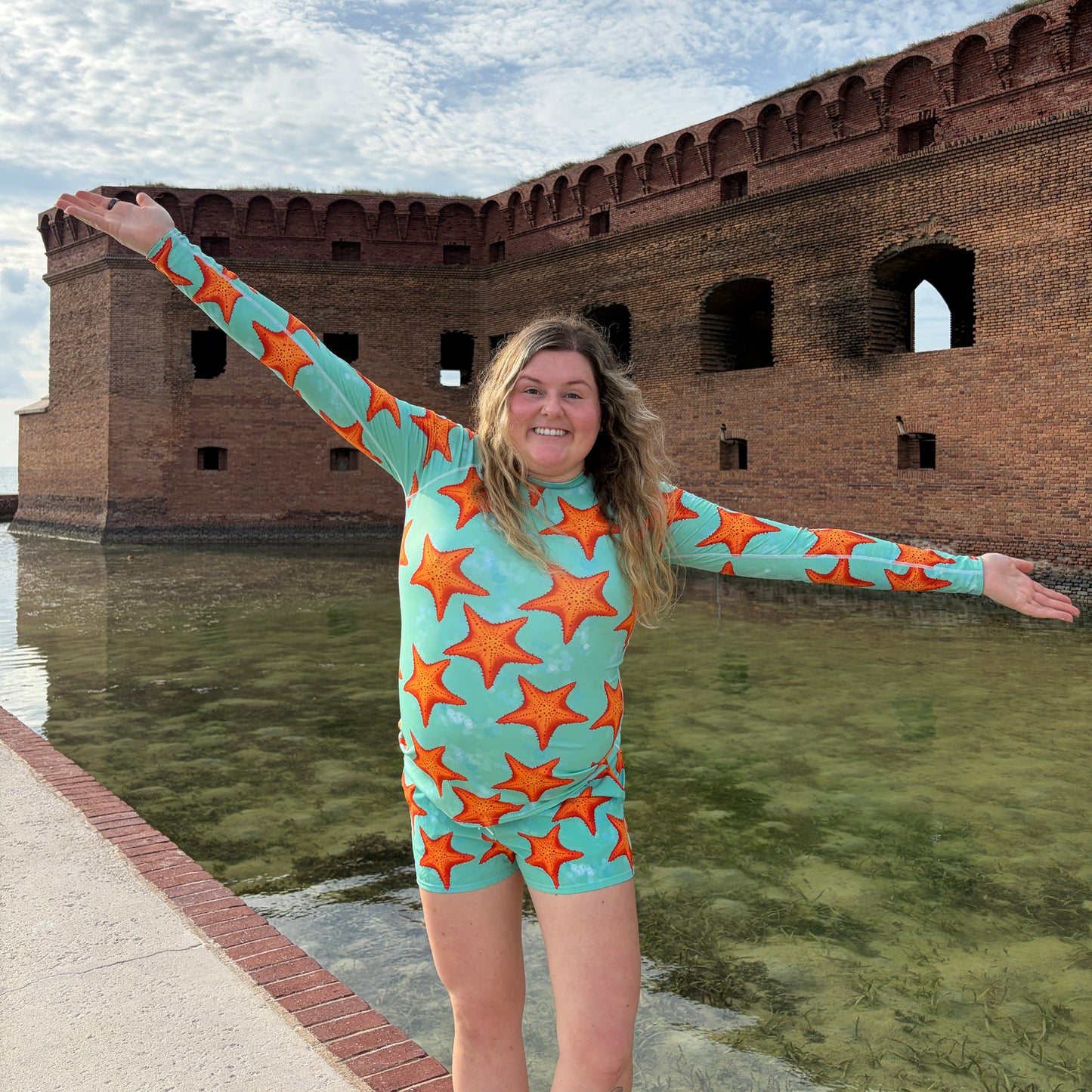 woman standing in front of water and a brick structure on a concrete path wearing a green/teal and orange starfish print rashguard and shorts 