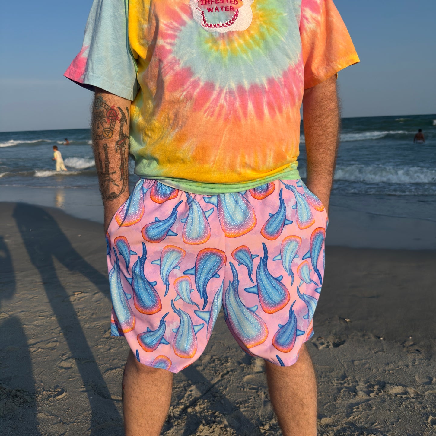 person standing on a sandy beach in front of water wearing pink colorful whale shark trunks and a tie dye shirt 