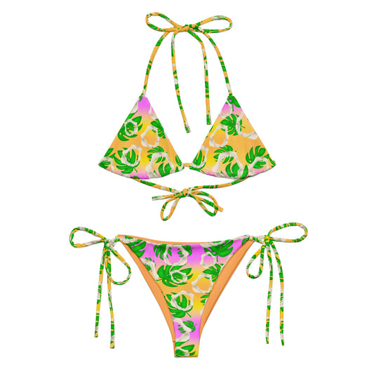A warm golden yellow and pinky lilac sunset ombré background with megalodon jaws and monstera leaves on our Meg Monstera Recycled string bikini 
