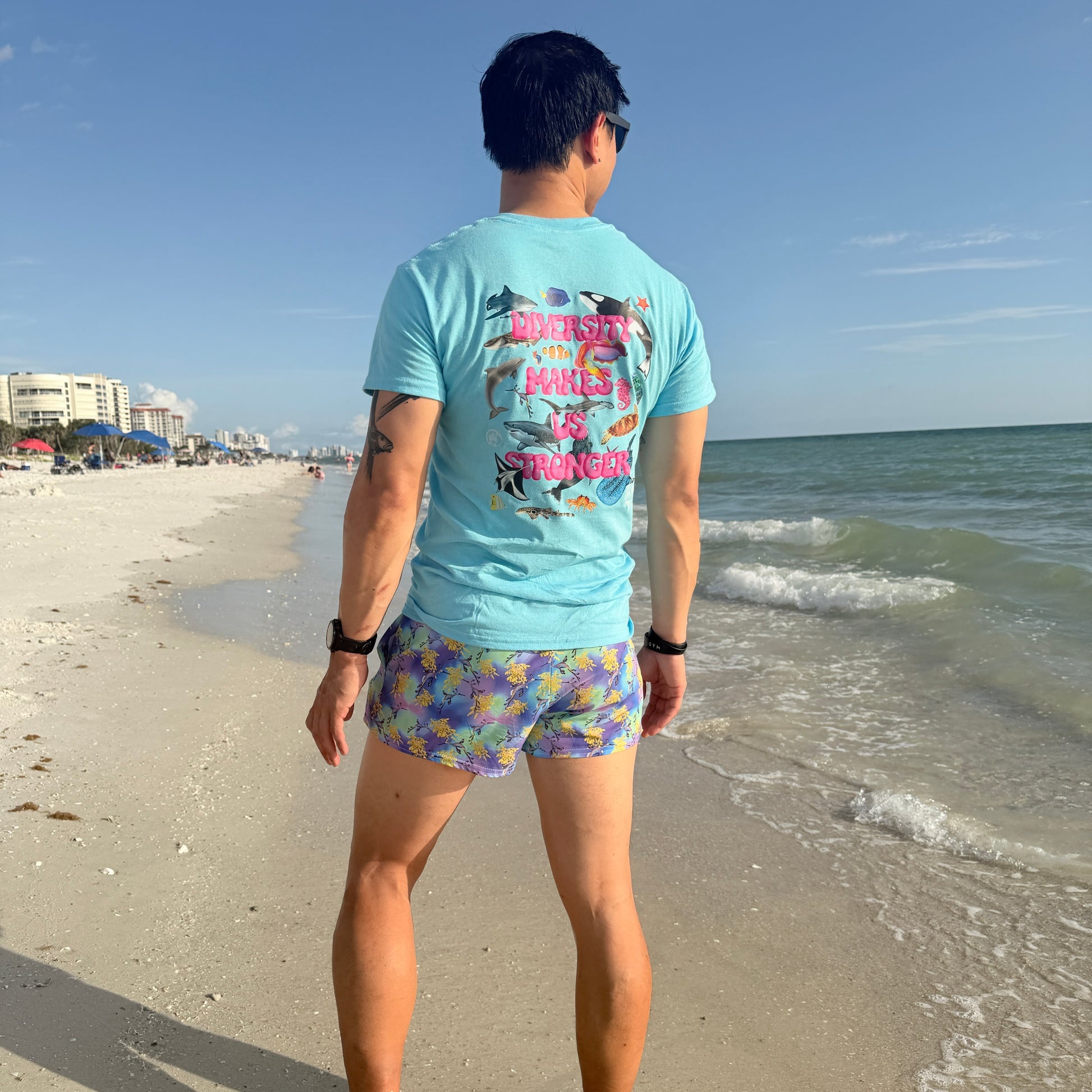 man standing on a beach facing the water wearing green, purple, yellow and blue swim shorts with sea dragons on them and a blue t shirt 