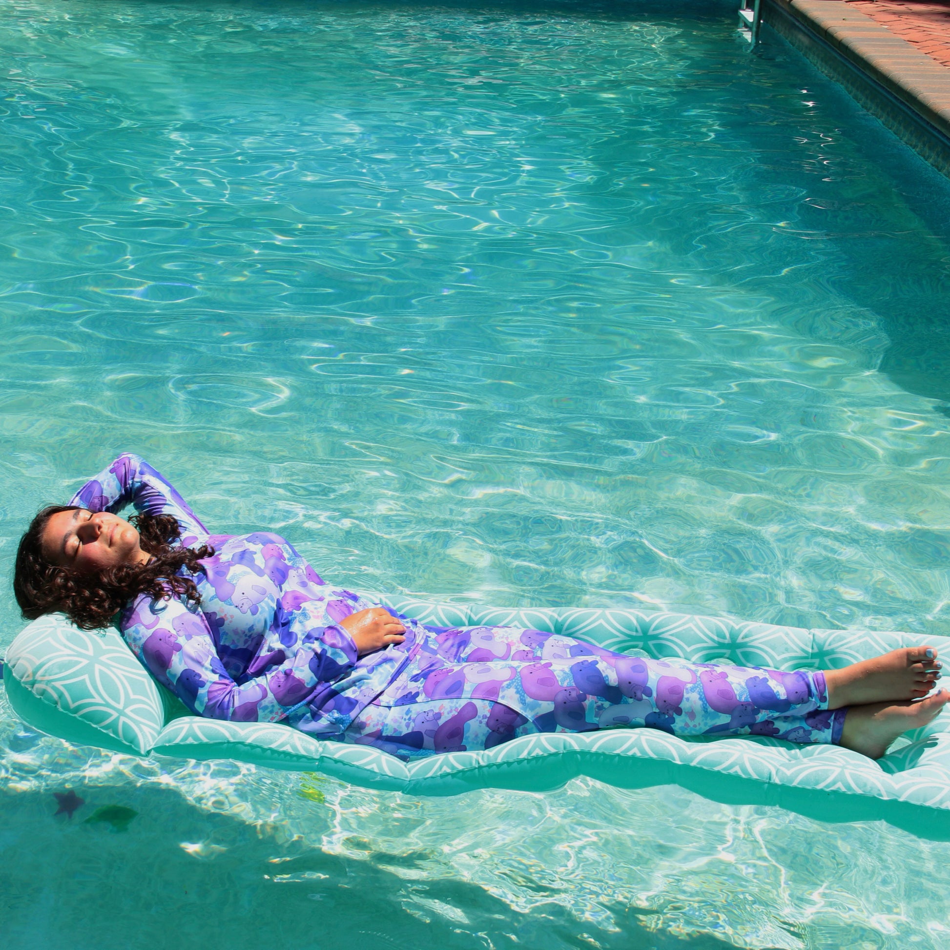 woman on a float laying in a swimming pool wearing a purple and blue rashguard and leggings with manatees on them 