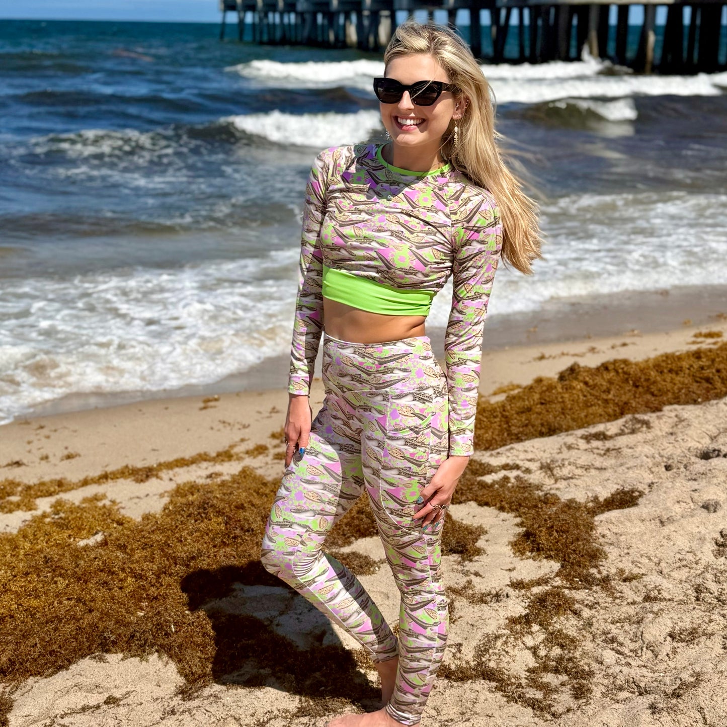 woman standing on the beach by the ocean wearing a green and pink cropped rashguard and leggings with zebra sharks on it. 