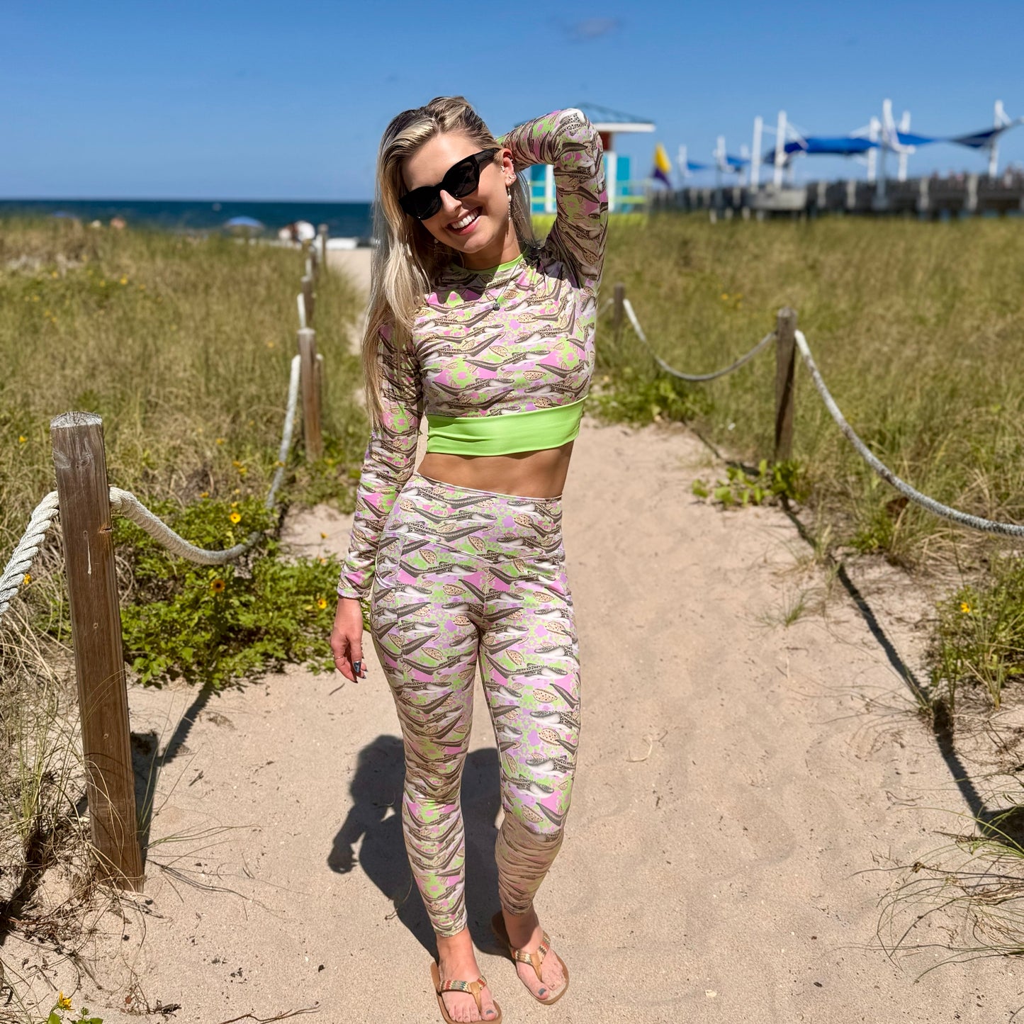 woman standing on a beach walkway by the ocean wearing a green and pink cropped rashguard and leggings with zebra sharks on it. 