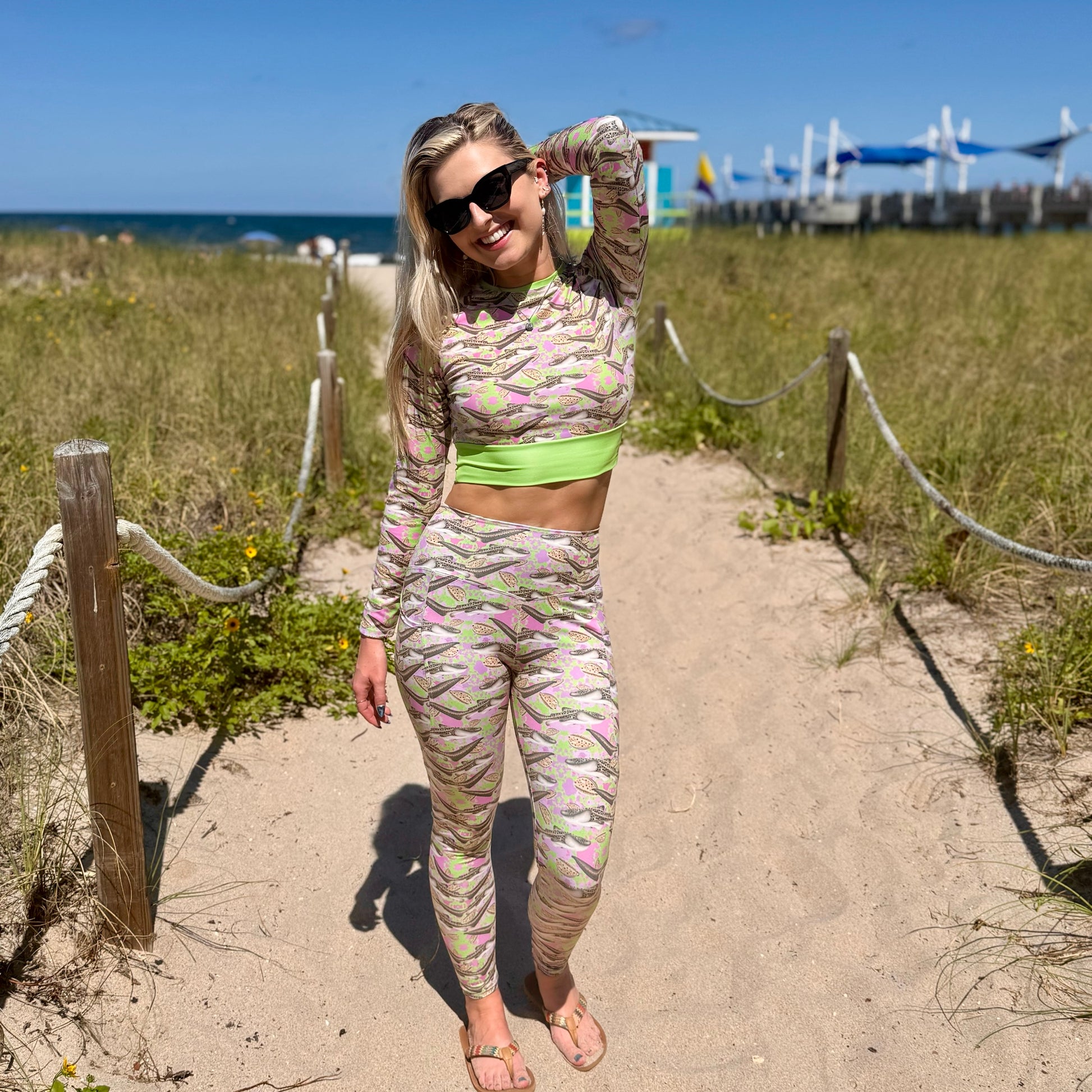 woman standing on a beach walkway by the ocean wearing a green and pink cropped rashguard and leggings with zebra sharks on it. 