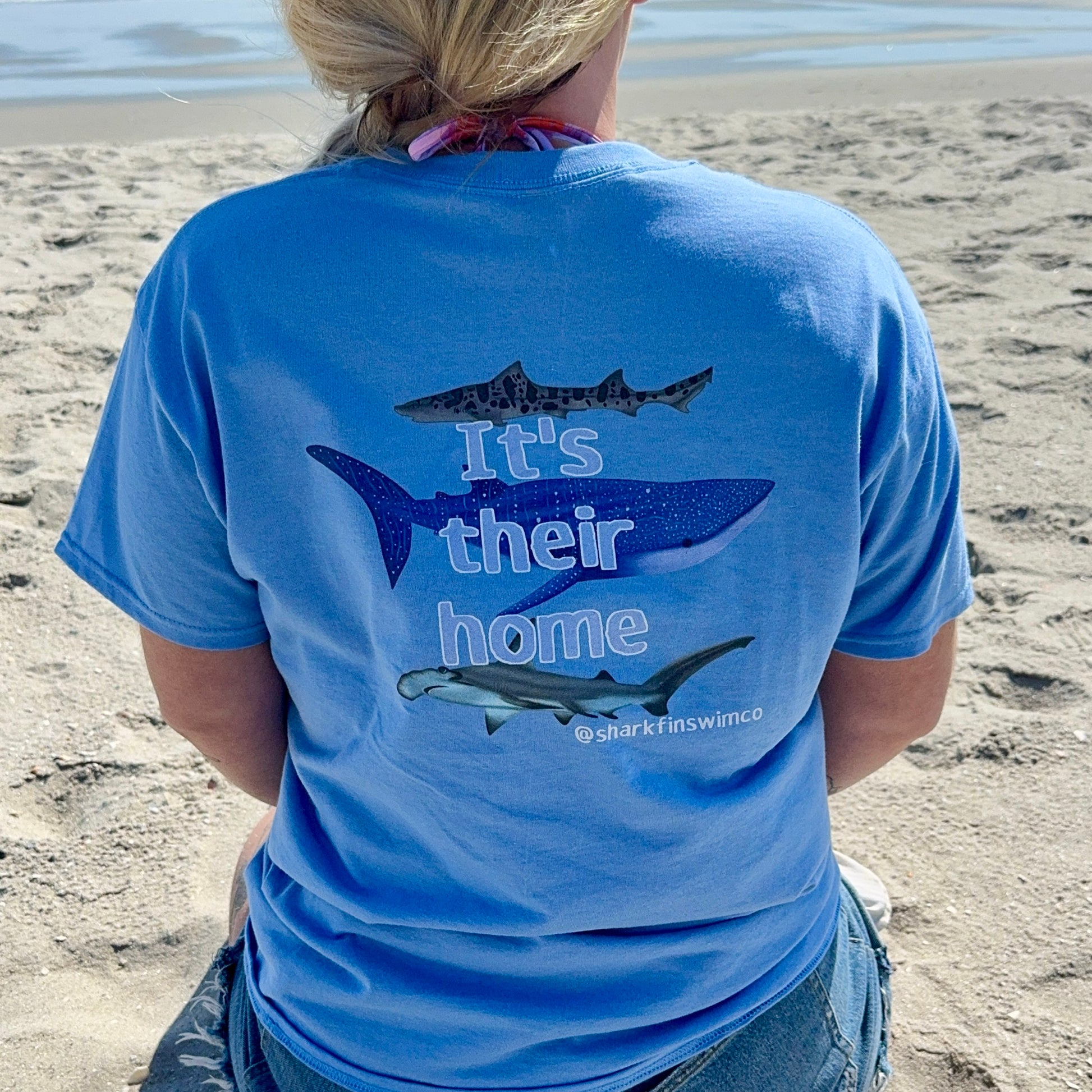 Person wearing a blue t-shirt with shark graphics and text on a beach