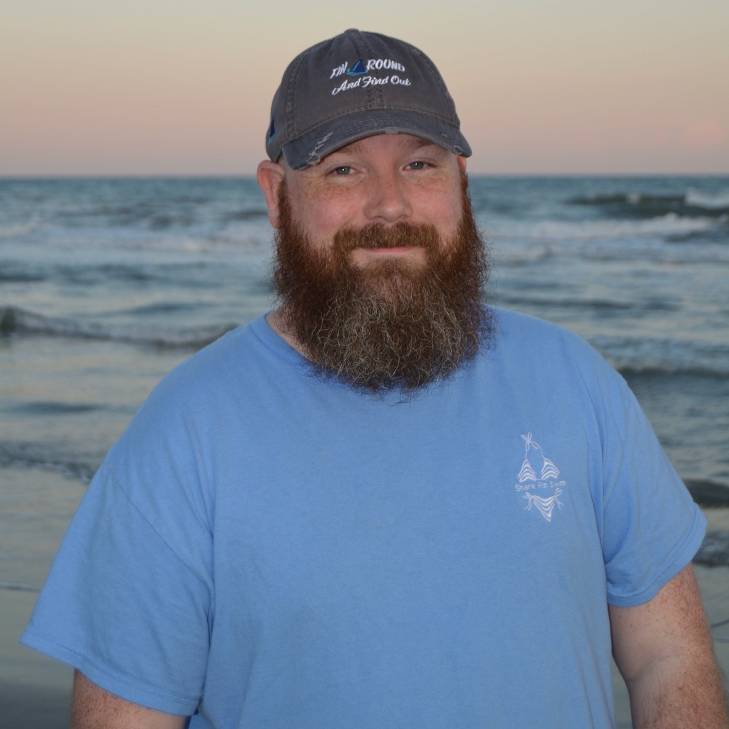 Man with a beard wearing a blue cap and shirt on a beach at sunset