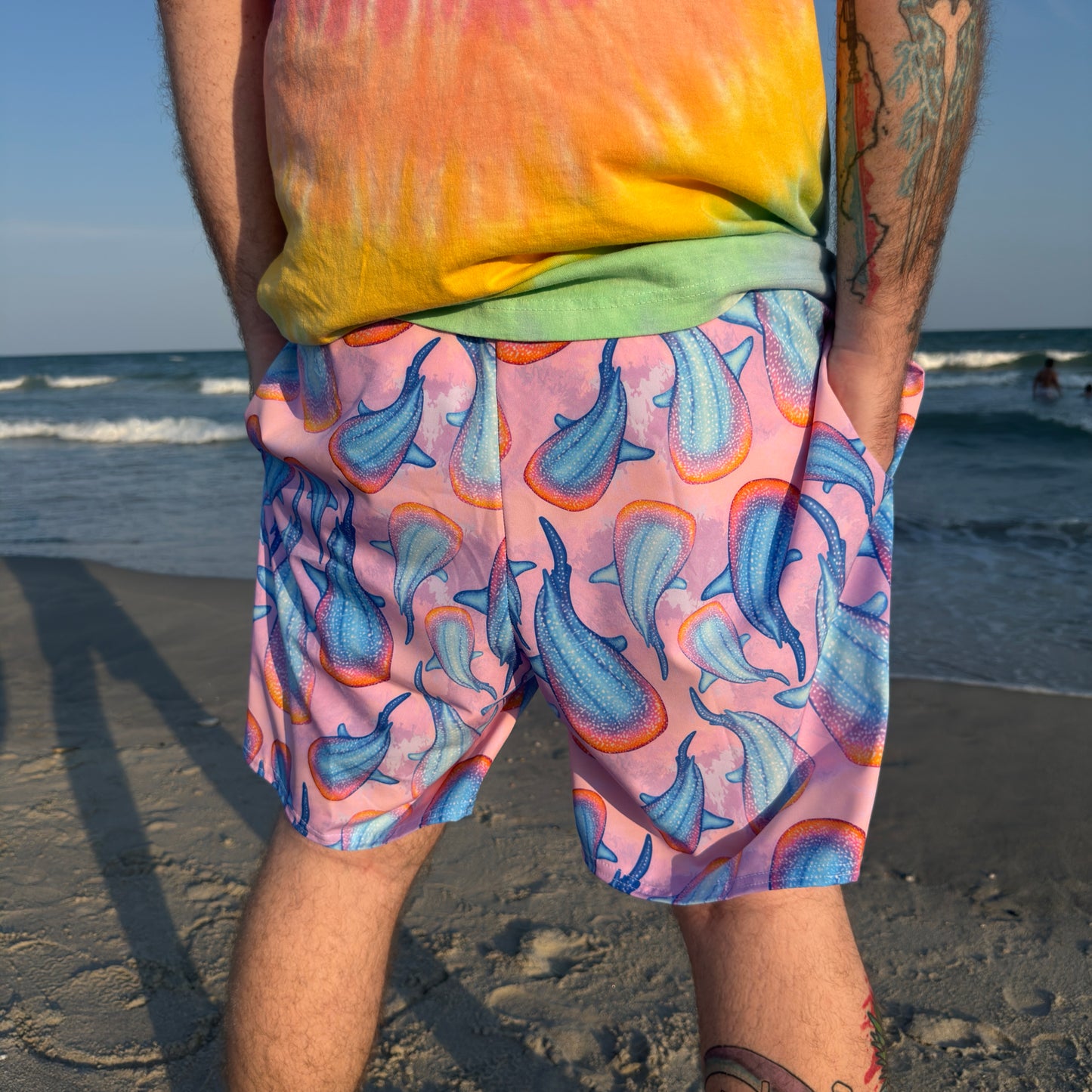 person standing on a sandy beach in front of water wearing pink colorful whale shark trunks and a tie dye shirt facing away from the camera 