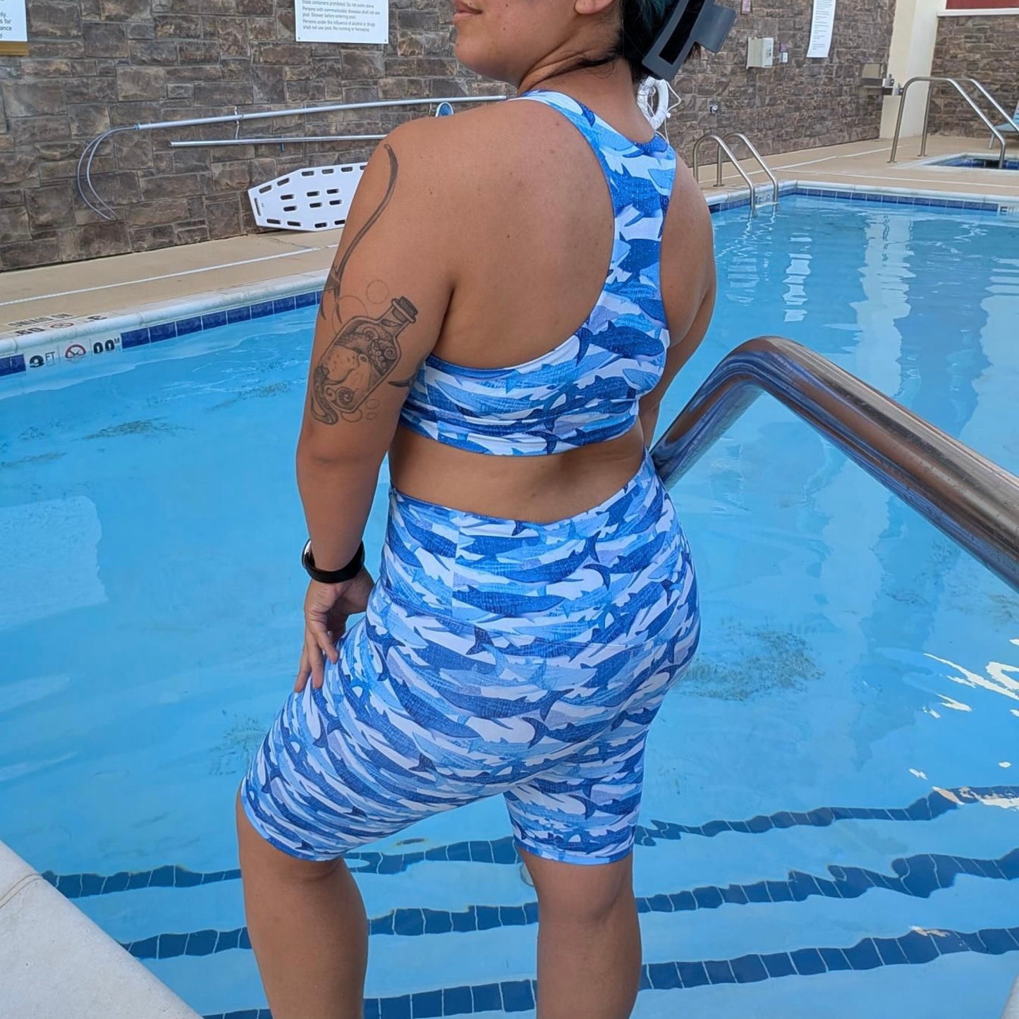 woman with black and blue hair standing in a pool facing to the side wearing blue whale shark sports bra and active shorts 