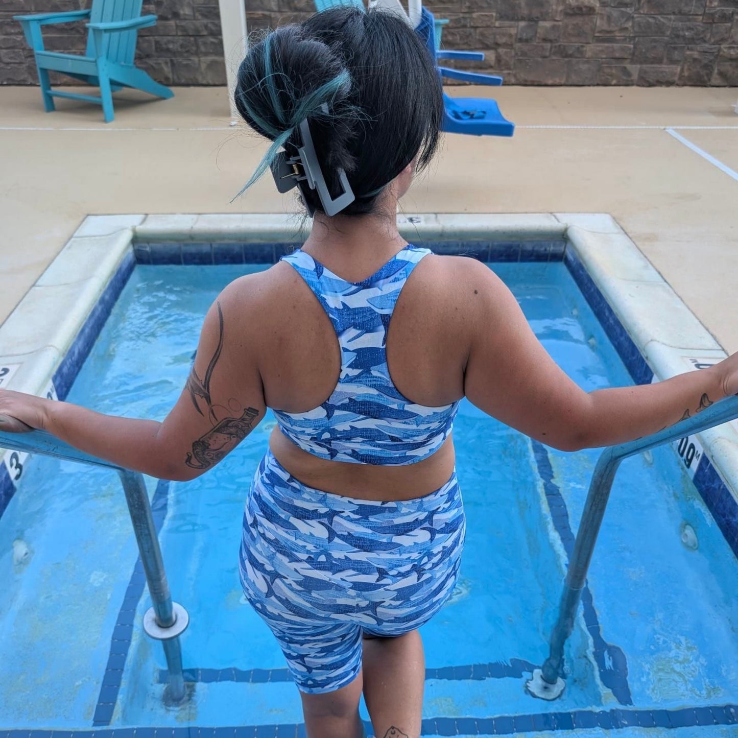 woman with black and blue hair wearing blue whale shark sports bra and active shorts standing in a pool facing away from the camera 