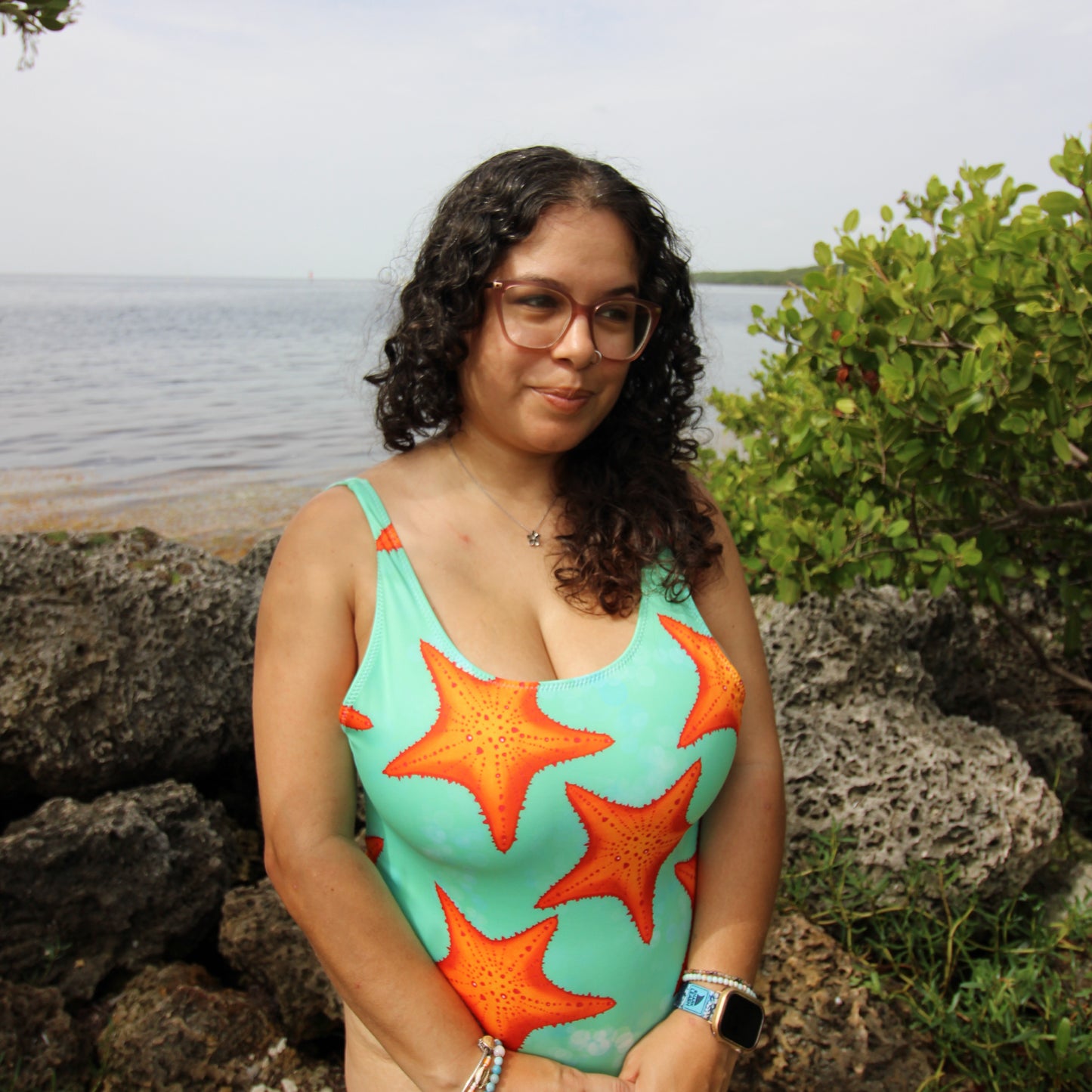 woman wearing a green and orange one piece starfish print swimsuit near water and rocks 