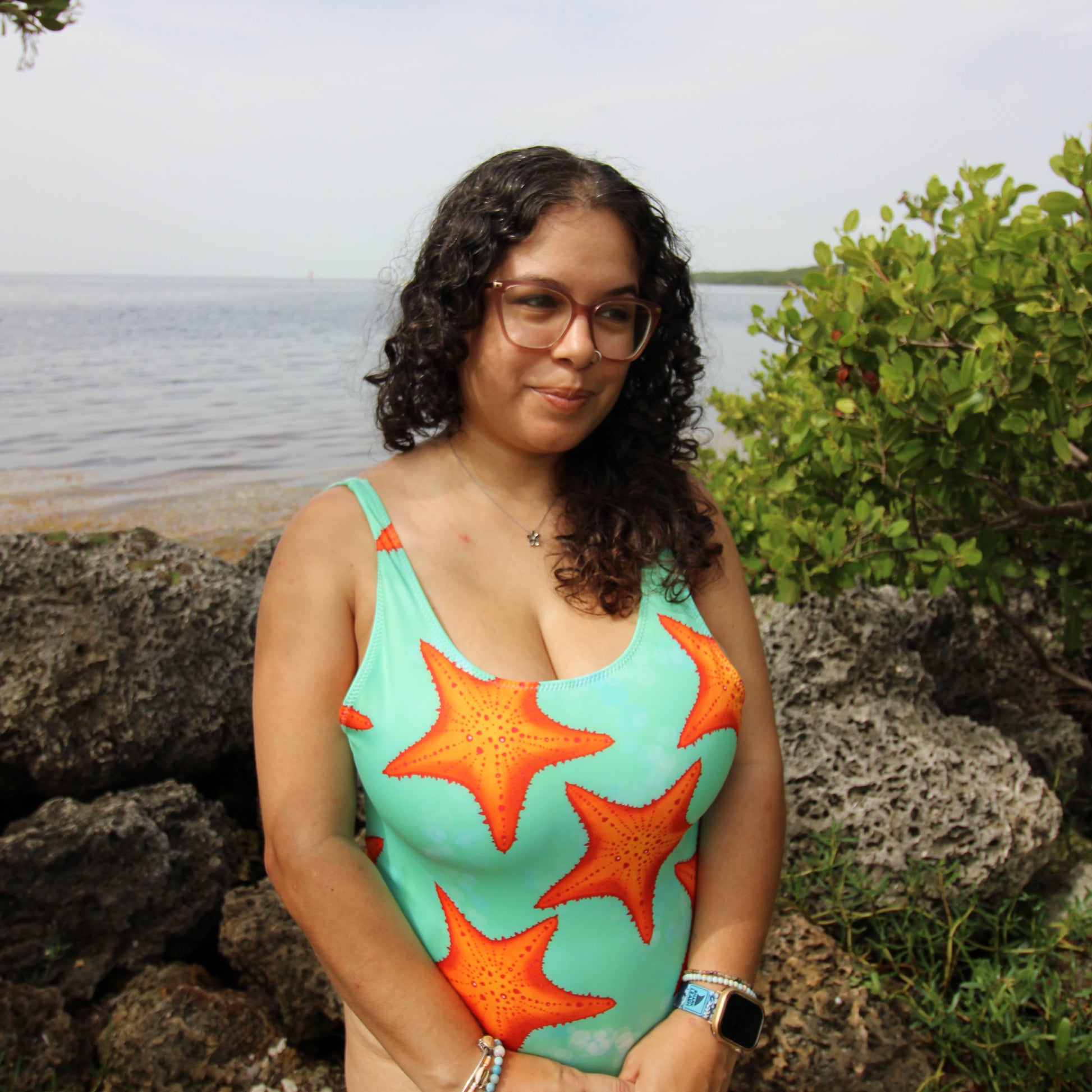 woman wearing a green and orange one piece starfish print swimsuit near water and rocks 
