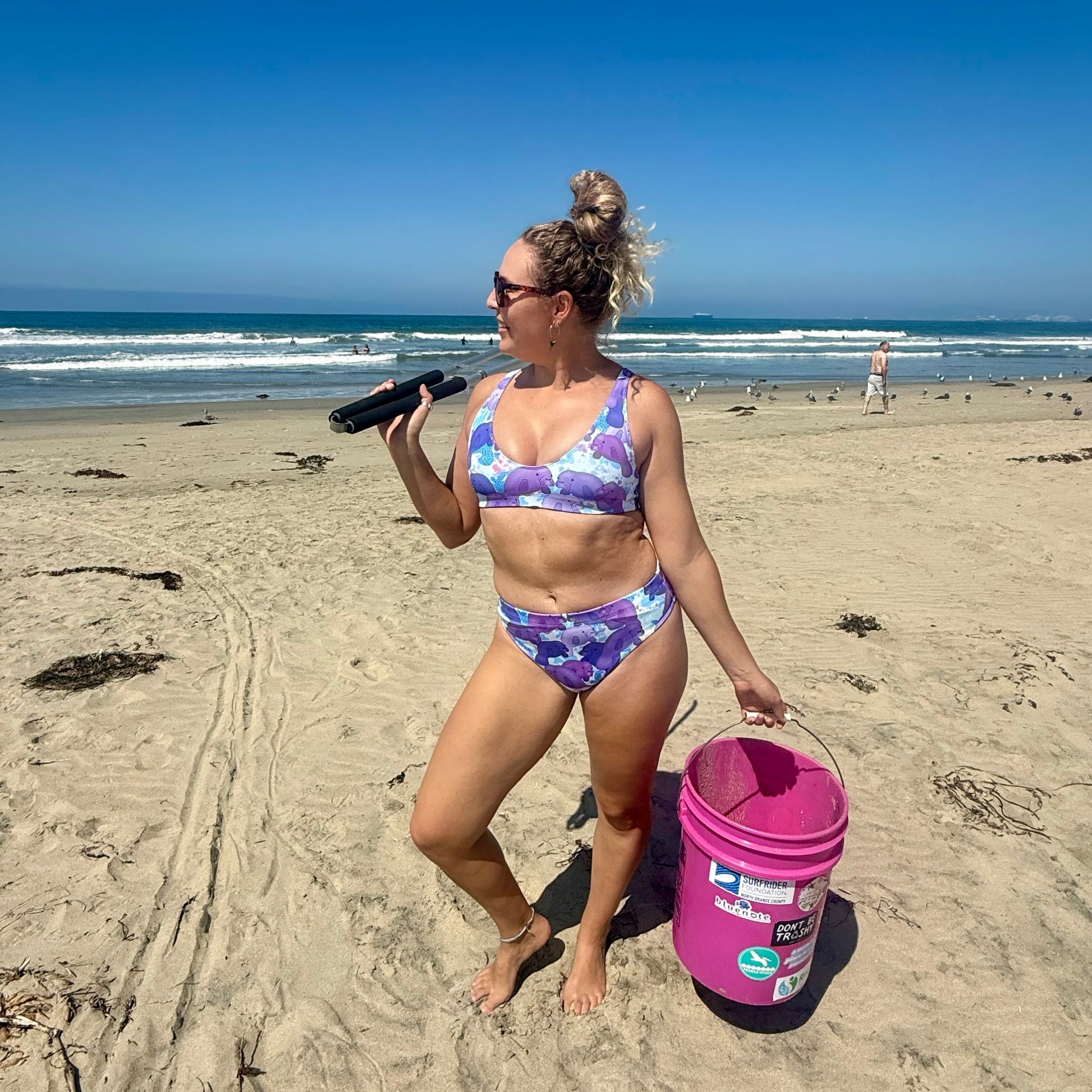 woman wearing a purple and pink manatee bikini on a beach with a litter bucket and tongs 
