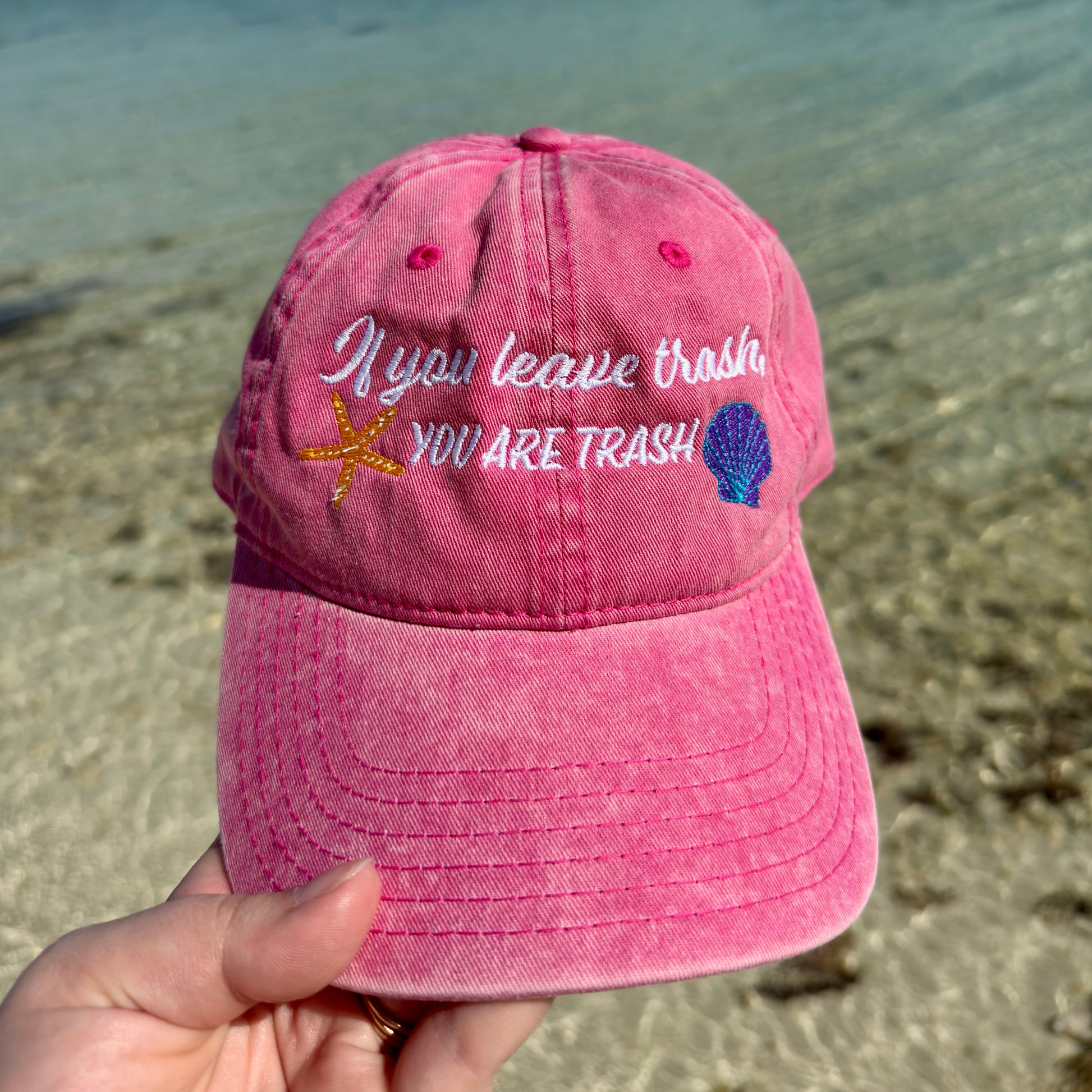 Pink baseball cap with embroidered ocean animals and text “if you leave trash, you are trash” In front of water 