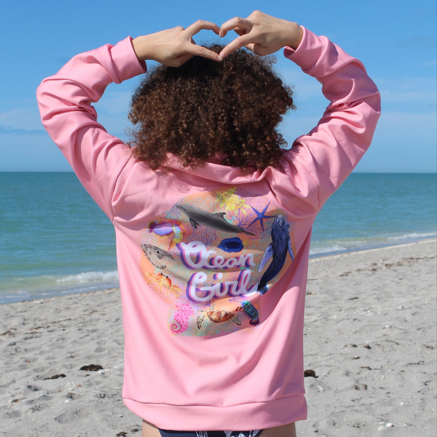 Woman standing on the beach wearing the ocean girl crew neck sweatshirt making a heart with her hands 