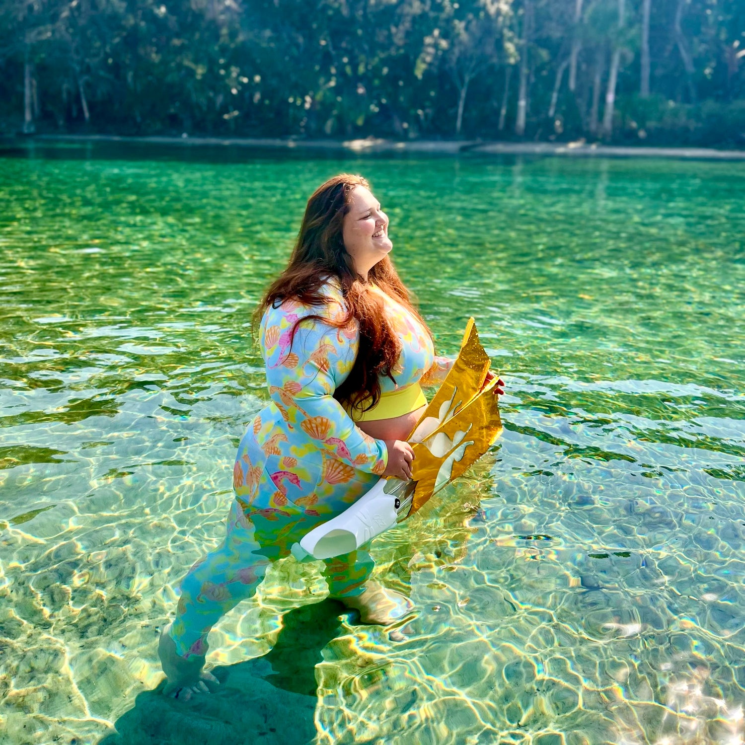 Aurora wearing Pink and orange seahorses with sunrise seashells. Light aqua blue and yellow background with a neon yellow band. Eco dive leggings and cropped rashguard with gold free diving fins in the water 