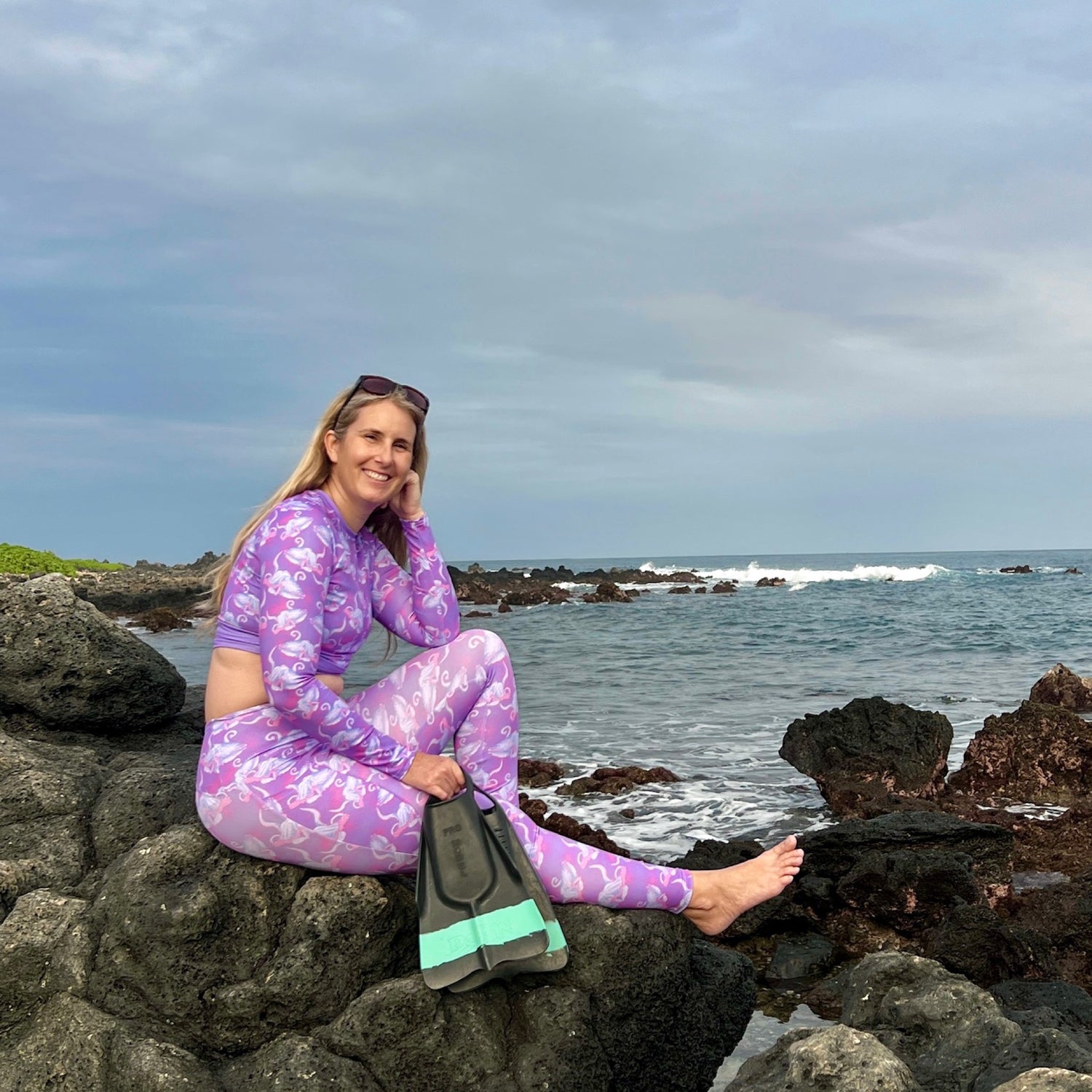 Woman sitting on a rocky beach wearing a cropped long sleeve rashguard in purple and leggings with swim fins 