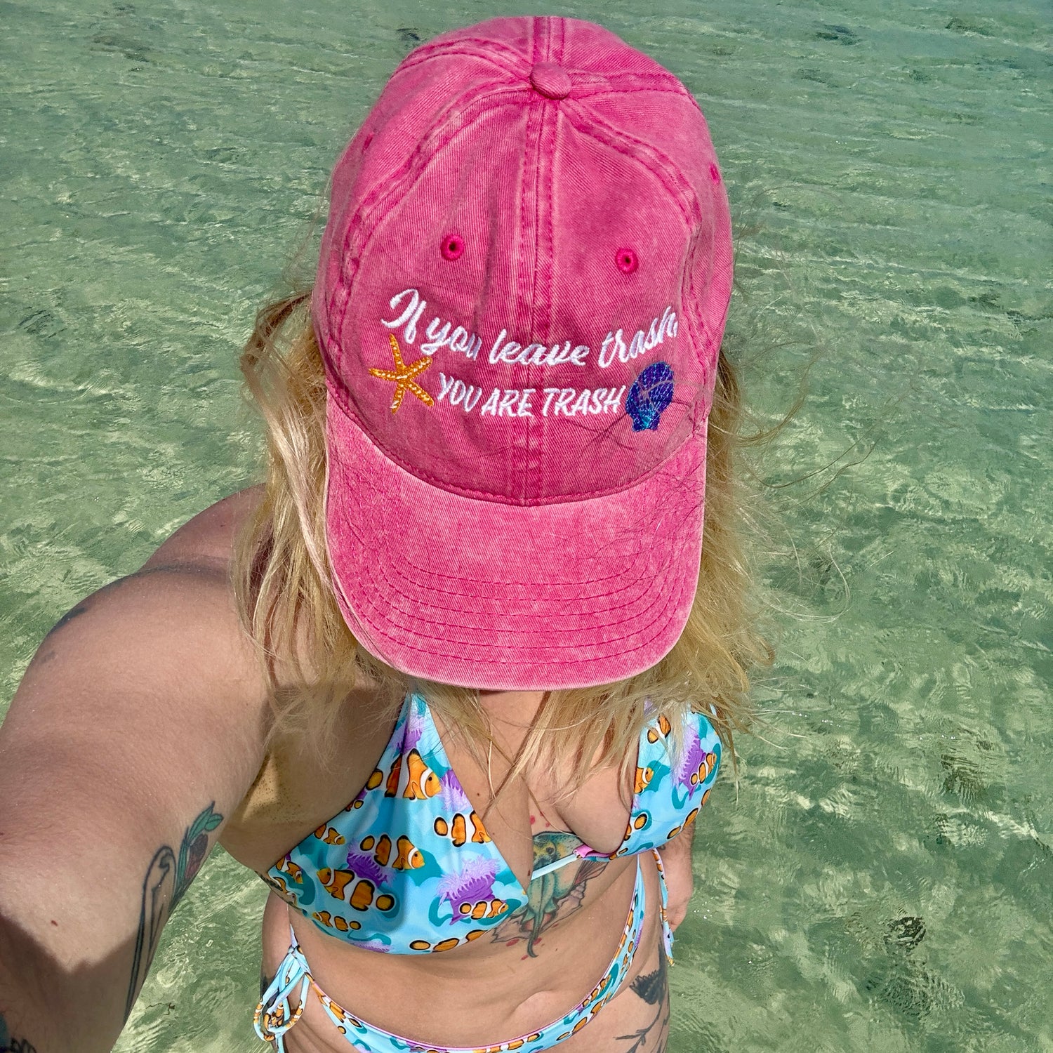 Woman standing in water wearing a pink ball cap with colorful ocean animals that says ”if you leave trash, you are trash” and a blue and pink clownfish bikini 